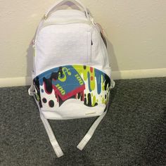 a white backpack with colorful designs on the front and back straps, sitting against a wall