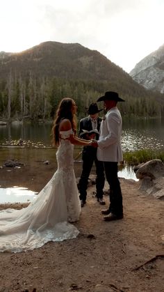 a bride and groom standing next to each other in front of a mountain lake holding hands