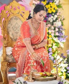 a woman in an orange sari sitting on a gold chair with flowers behind her