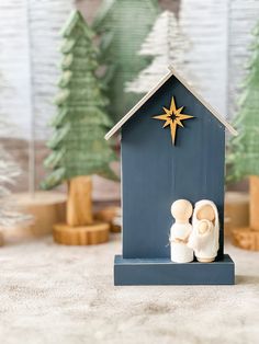 a nativity scene with two wooden figures in front of a blue house and christmas trees