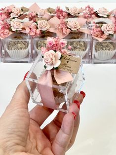 a hand holding a small glass box with flowers in it next to some cupcakes