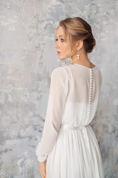 a woman wearing a white dress with pearls on the back and long sleeves, standing in front of a gray wall