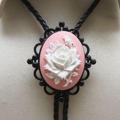 This cool very different handmade  bolo tie features a white rose on a pink resin cameo as the slide.   It is a nicely piece measuring 2 1/4  inches tall by 2 inches wide. There is a black metal Gothic setting around the cameo .  It is in excellent condition and mounted upon a  36 inch long black braided leatherette string tie with bright silverplated metal end caps.    This was handmade in our very own Montana Rock shop.  We will send this to you in a lovely gift box for safe storage or easy gi Gothic Setting, Bolo Ties, Safe Storage, Bolo Tie, Black Braids, Tie Accessories, Suit And Tie, Vintage Rhinestone, Easy Gifts