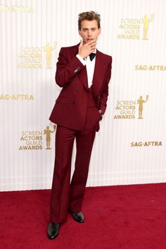 a young man in a red suit and bow tie standing on a red carpet with his hand to his face