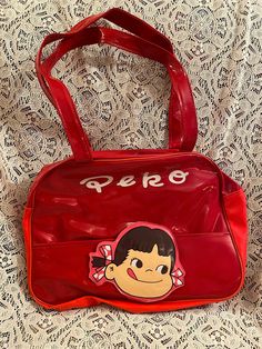 This sweet Peko tote has a shiny red look. The cute little girl head on the front of the tote has velcro  and can be opened. It zips open at the top. It has a zipper in the back as well. We have added a flower charm to the zipper as part of it has cracked off, thus making it easier to open and close. The inside of the bag has a little bit of light water staining as well.  It is an awesome tote and in overall nice vintage condition. Measurements: Length: 10 inches Width: 14 inches Strap Drop: 11 inches Retro Red Shoulder Bag With Zipper Closure, Playful Red Shoulder Bag For Daily Use, Retro Red Bag With Zipper Closure, Cute Red Satchel Bag, Retro Red School Shoulder Bag, Cute Red Shoulder Bag With Zipper Closure, Fun Red Tote Bag, Light Water, Late 90s