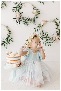 a baby girl in a blue dress eating cake with flowers on her head and greenery behind her