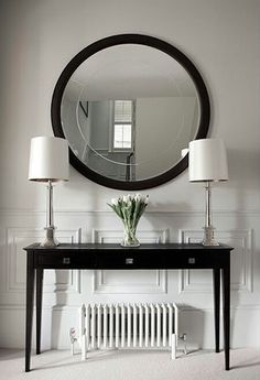 a black table with two lamps and a round mirror on the wall above it in a white room