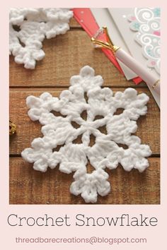 the crochet snowflake is laying on top of a wooden table next to a pen and paper