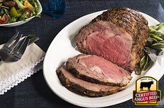 a white plate topped with sliced meat next to a bowl of salad and utensils