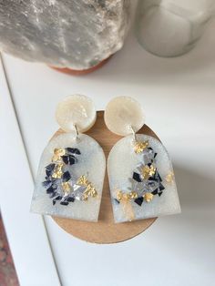 a pair of white and gold earrings sitting on top of a wooden board next to a rock