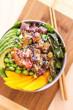 a bowl filled with vegetables and meat on top of a wooden cutting board next to chopsticks