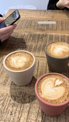 three cups of coffee sitting next to each other on top of a wooden table with two cell phones