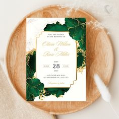 an elegant wedding card with green leaves and gold foil is on top of a wooden plate