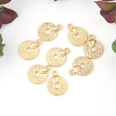 gold plated charms with circular design on white surface next to flowers and leaves in the background