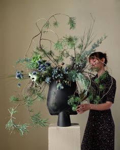 a woman standing next to a black vase filled with green and blue flowers on top of a white pedestal