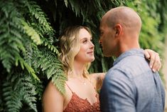 a man and woman standing next to each other in front of green plants, looking at each other