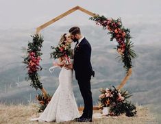 a bride and groom kissing in front of an arch decorated with floral wreaths on top of a mountain