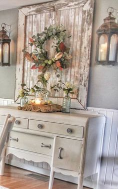 a white dresser sitting next to a wooden table with flowers on it and two lanterns hanging from the wall