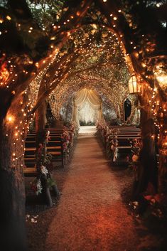 A whimsical wedding aisle under an archway of trees adorned with twinkling fairy lights. Wooden benches line the path, which leads to a draped, romantic altar. Floral arrangements decorate the seating area, creating an enchanting forest wedding setting. Wedding Under Tree With Lights, Elf Themed Wedding, Indoor Enchanted Forest Wedding, Magical Forest Wedding Theme, Whimsical Wedding Aisle, Fairy Wedding Aesthetic, Mystical Wedding Theme, Altar Floral Arrangements, Enchanted Wedding Themes