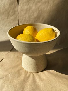 three lemons in a white bowl sitting on a beige cloth covered tablecloth,