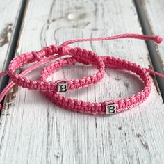 two pink braided bracelets with black letters on them sitting on a white wooden surface