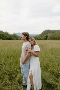 a young man and woman standing in tall grass with their arms around each other, looking into the distance