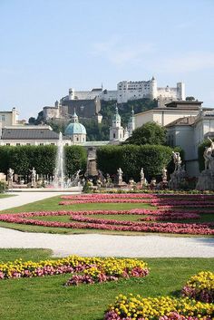 an elaborate garden with flowers in front of a large building and water fountain on the other side