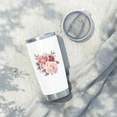 a white tumbler with pink roses on it next to a can of water and a blanket