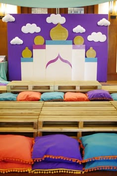 colorful pillows are arranged on wooden benches in front of a purple wall with clouds above it