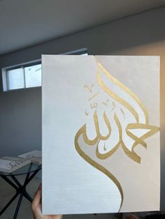 a hand holding up a white and gold painting with the word in arabic on it