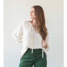 a woman standing in front of a white wall wearing green pants and a white blouse