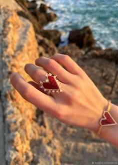 a woman's hand with a red and white ring on it next to the ocean