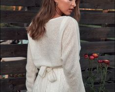 a woman standing in front of a wooden fence wearing a white sweater and black pants