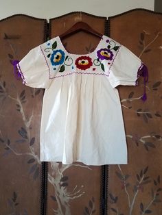 80s lovely ivory cotton Mexican hippie boho peasant blouse. Purple red and blue embroidered flowers with swirly green leaves. The square neck is edged in bright purple so are the cuffs of sleeves with long purple tassels Pleated front and back. Bust small 36 Across shoulders 13 Length 26 Spring Festival Beige Peasant Top, Bohemian Blouse With Embroidered Sleeves, Bohemian Summer Peasant Top With Embroidered Sleeves, White Hippie Peasant Top For Festival, Bohemian Peasant Top With Embroidered Sleeves For Summer, Fall Cotton Short Sleeve Peasant Top, Fall Cotton Peasant Top With Short Sleeves, Fall Short Sleeve Cotton Peasant Top, Summer Festival Peasant Top With Embroidered Sleeves