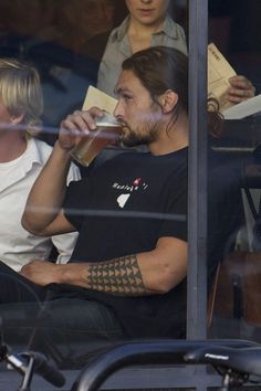 a man with tattoos drinking from a glass while sitting in front of a window next to a woman