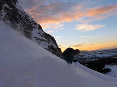 a person on skis going down a snowy hill at sunset or dawn with mountains in the background