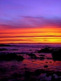 the sun is setting over the ocean with rocks in the foreground and purple sky