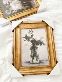 two framed pictures with flowers in them sitting on a white bed sheet, next to each other