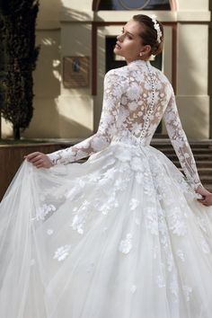 a woman in a white wedding dress with flowers on the skirt and long sleeves is standing outside