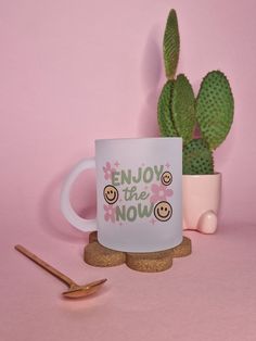 a white coffee mug with the words enjoy the now printed on it next to a cactus