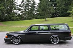 an old black station wagon parked in a parking lot next to some trees and grass