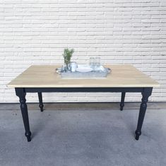 a wooden table with two black legs and a glass vase on top, against a white brick wall