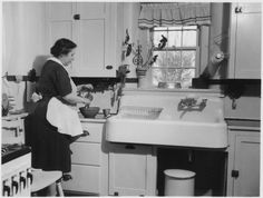 an old black and white photo of a woman washing dishes in the kitchen with her dog