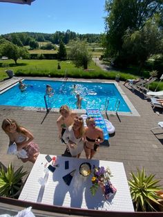 three women sitting at a table in front of a swimming pool with people standing around it