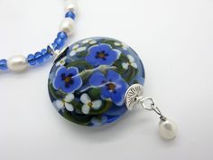 A long necklace with a large handmade flower bead. It is made of a transparent blue glass and decorated decorated with white and blue flowers. Under the large lentil pearl is a movable freshwater cultured pearl. The necklace is threaded with blue transparent glass rocailles  and freshwater cultured pearls. It is closed with a lobster clasp. The circulating length is about 90 cm. In addition, the pendant unit, this measures about 6.5 cm. This necklace goes very well with plain clothes.  Single pi Blue Flower Shaped Beaded Necklace For Gift, Blue Flower-shaped Beaded Necklace Gift, Handmade Blue Flower Pendant Necklace, Plain Clothes, Painting On Glass, Bead Flower, Lampwork Jewelry, White And Blue Flowers, Plain Outfits