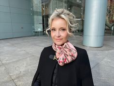 a woman with blonde hair wearing a black jacket and pink scarf standing in front of a building