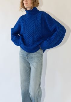 a woman standing in front of a white wall wearing a blue sweater and denim jeans