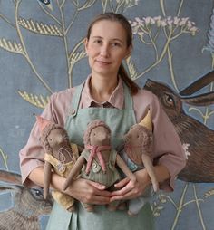 a woman holding three stuffed animals in front of a wall with birds and flowers on it