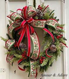 a christmas wreath with red and green ribbons hanging on a white front door, decorated with pine cones