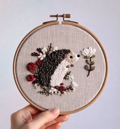 a hand holding up a small embroidered hedge with flowers on it's back and behind it is an embroidery hoop